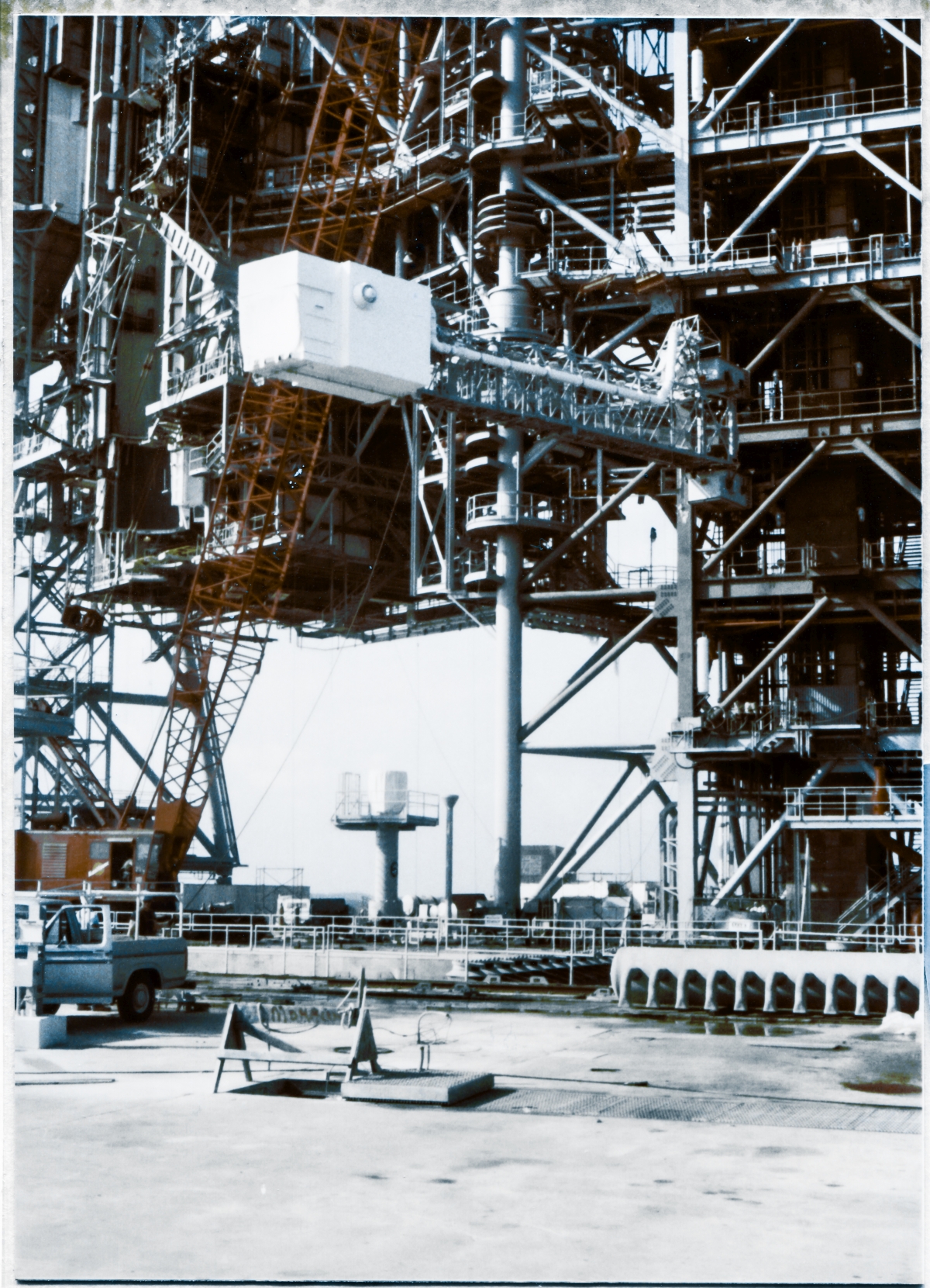 Image 133. OAA Lift 5 of 9. Against a bewildering background of structural steel elements making up the RSS and the FSS at Space Shuttle Launch Complex 39-B, Kennedy Space Center, Florida, the Orbiter Access Arm (OAA) being lifted by the large red Manitowoc crane, rises toward its final location out of view above the top margin of this frame, where it will be bolted onto its support Strongback at Elevation 200'-0” by Union Ironworkers working for Ivey Steel Erectors. The OAA is what the crew walked across to gain access to the Hatch in the side of the Space Shuttle Orbiter. The opening in the white Environmental Chamber at the left end of the Arm which the crew would pass through, has been covered in order to keep its interior clean and free of external contaminants that might enter it, otherwise.  Photo by James MacLaren.